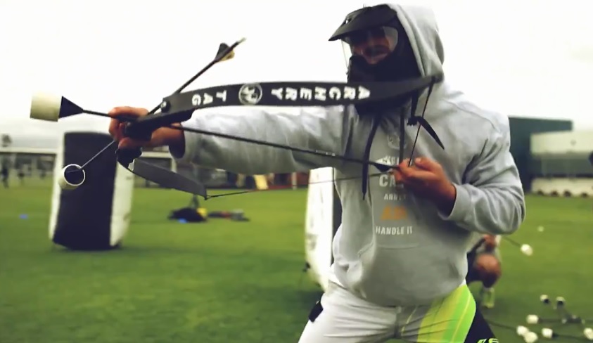 Joueur en sweat à capuche visant avec un arc d'Archery Tag, prêt à lancer une flèche, sur un terrain de jeu extérieur.